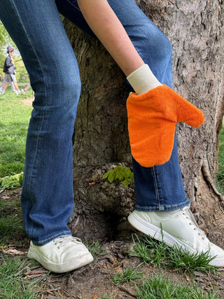 Person wearing blue jeans and white sneakers with an orange TiCK MiTT glove on a tree trunk.