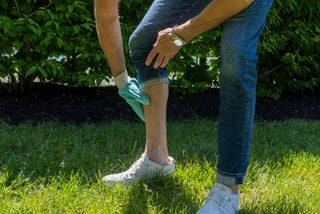 Person checking their lower leg for ticks while standing outdoors on grass.